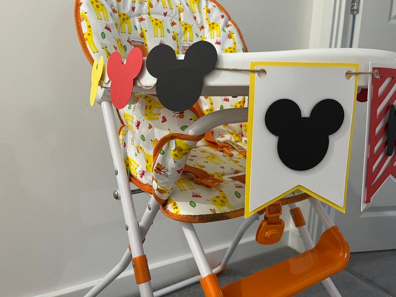 May include: A high chair with a giraffe-patterned seat and orange accents. A banner with Mickey Mouse head cutouts and pennants hangs across the front. The chair is white with orange and yellow details.