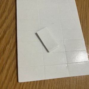 May include: A sheet of white, square adhesive labels with one label peeled off. The labels are arranged in a grid pattern. The background is a wooden surface.