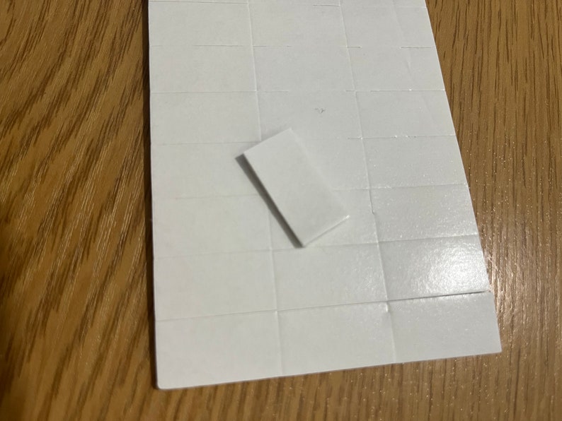 May include: A sheet of white, square adhesive labels with one label peeled off. The labels are arranged in a grid pattern. The background is a wooden surface.