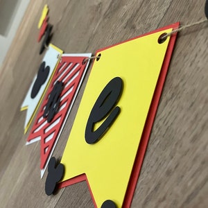 May include: A colourful banner with the letter "e" in black, displayed on a wooden surface. The banner features yellow, red, and white pennants with Mickey Mouse-themed accents. The banner is strung together with twine.