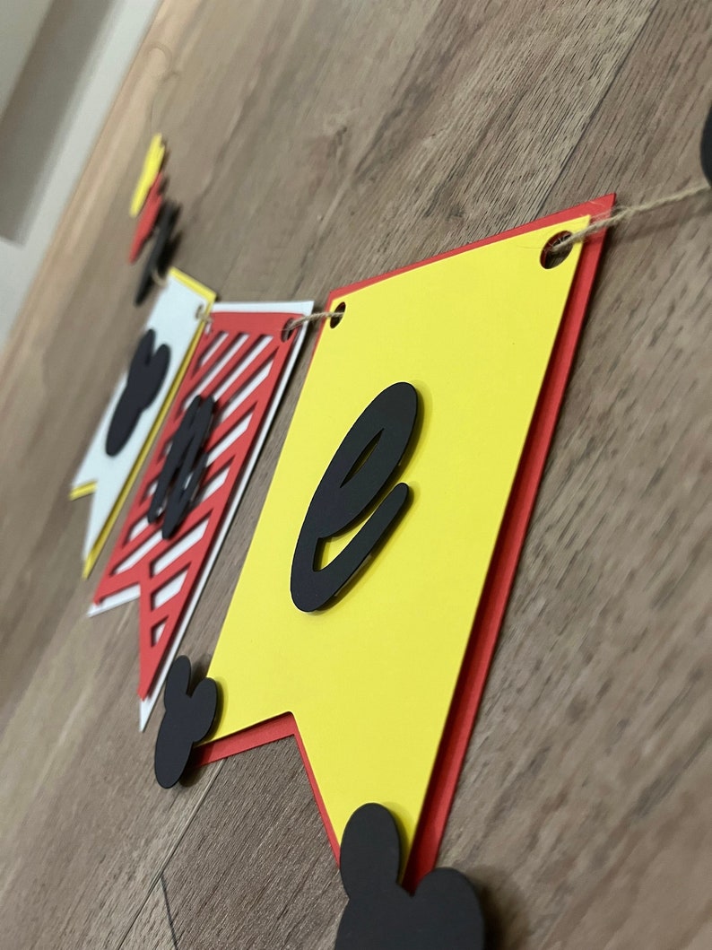 May include: A colourful banner with the letter "e" in black, displayed on a wooden surface. The banner features yellow, red, and white pennants with Mickey Mouse-themed accents. The banner is strung together with twine.