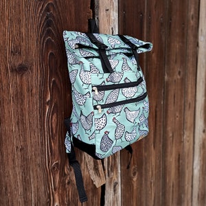 Op de afbeelding: Een lichtblauwe rugzak met een herhaald patroon van zwart-witte kippen. De rugzak heeft drie vakken met ritssluiting, zwarte bandjes en een oprolbare sluiting met zwarte gespen. De tas hangt aan een houten oppervlak.