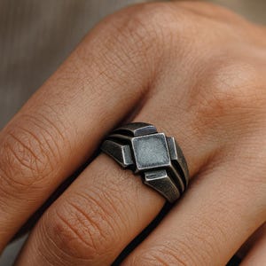 May include: A silver-toned ring with a geometric design featuring a square center and stepped details. The ring is worn on a finger, showcasing its intricate, industrial-inspired style.