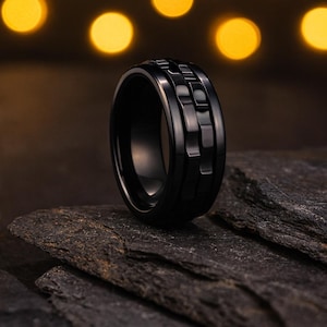 Op de afbeelding: Een zwarte ring met een getextureerd, geometrisch ontwerp. De ring is gemaakt van een donker, glanzend materiaal en ligt op een donker, getextureerd oppervlak. De achtergrond heeft vage gele lichten, wat een warm contrast creëert.