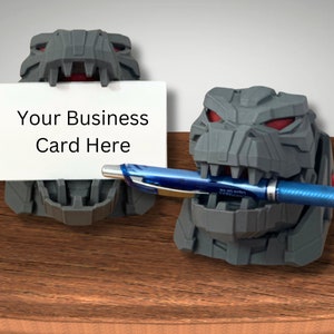 May include: Gray 3D printed dinosaur head desk organizer with red eyes. The dinosaur is holding a blue pen in its mouth and a white business card that says "Your Business Card Here".