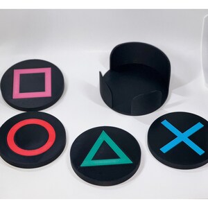 May include: Set of four black coasters with different colored symbols: a pink square, a red circle, a green triangle, and a blue X. The coasters are made of a soft, rubbery material and are perfect for protecting surfaces from scratches and spills.