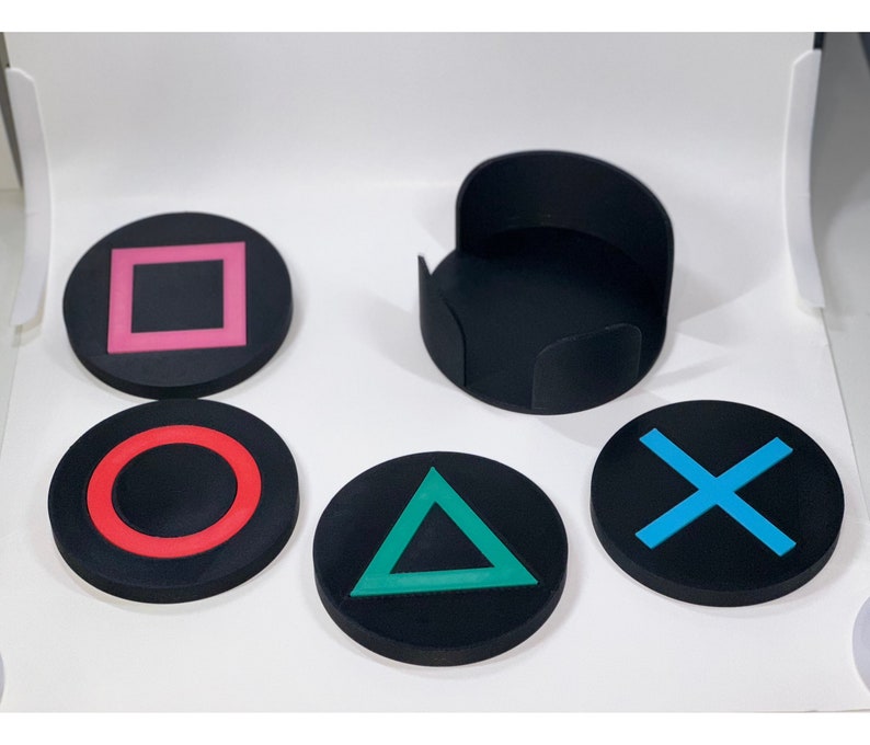 May include: Set of four black coasters with different colored symbols: a pink square, a red circle, a green triangle, and a blue X. The coasters are made of a soft, rubbery material and are perfect for protecting surfaces from scratches and spills.