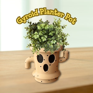 May include: A brown ceramic planter pot shaped like a Gyroid from the Animal Crossing video game series. The pot has a face with two black eyes and a mouth, and is decorated with small black dots. The pot is filled with a green leafy plant.