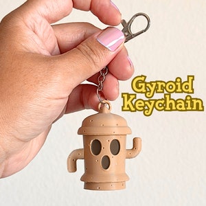 May include: A brown Gyroid keychain with a silver chain and a silver clasp. The keychain is shaped like a small house with a face and a small tree growing out of the top.
