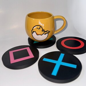 May include: Four black coasters with different designs. One coaster has a square, one has a plus sign, one has a circle, and one has a red circle. A yellow mug with a cartoon character and the text "meh..." is sitting on top of the coaster with the red circle.