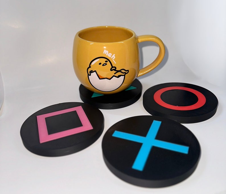 May include: Four black coasters with different designs. One coaster has a square, one has a plus sign, one has a circle, and one has a red circle. A yellow mug with a cartoon character and the text "meh..." is sitting on top of the coaster with the red circle.