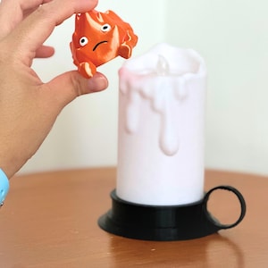 K&ouml;nnte beinhalten: Eine wei&szlig;e Kerze mit schwarzem Sockel und Griff. Die Kerze tropft Wachs und hat ein kleines Licht im Inneren. Eine kleine orangefarbene Figur mit einem traurigen Gesicht wird &uuml;ber der Kerze gehalten.