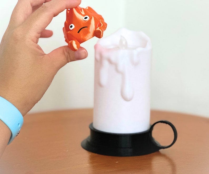 K&ouml;nnte beinhalten: Eine wei&szlig;e Kerze mit schwarzem Sockel und Griff. Die Kerze tropft Wachs und hat ein kleines Licht im Inneren. Eine kleine orangefarbene Figur mit einem traurigen Gesicht wird &uuml;ber der Kerze gehalten.