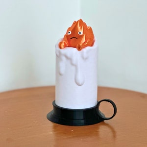 K&ouml;nnte beinhalten: Eine wei&szlig;e Kerze mit schwarzem Sockel und Griff. Die Kerze hat einen Effekt von geschmolzenem Wachstropfen. Die Oberseite der Kerze zeigt eine kleine orangefarbene Figur eines Feuergeistes mit einem traurigen Ausdruck.