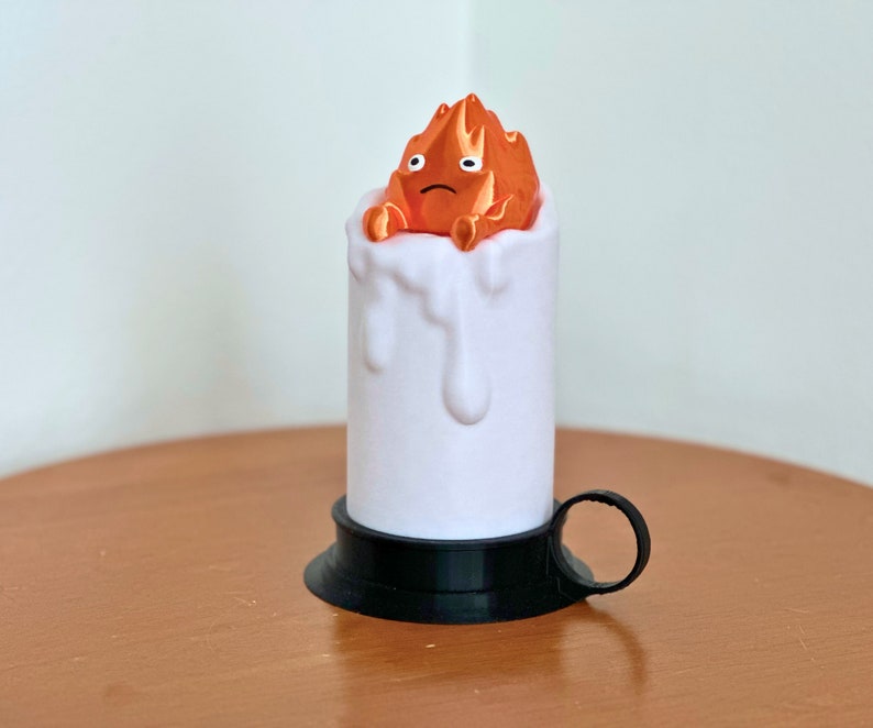 K&ouml;nnte beinhalten: Eine wei&szlig;e Kerze mit schwarzem Sockel und Griff. Die Kerze hat einen Effekt von geschmolzenem Wachstropfen. Die Oberseite der Kerze zeigt eine kleine orangefarbene Figur eines Feuergeistes mit einem traurigen Ausdruck.