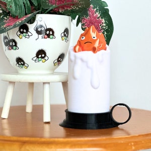 K&ouml;nnte beinhalten: Eine wei&szlig;e Kerze mit schwarzem Sockel und Griff. Die Kerze hat einen geschmolzenen Wachseffekt und eine kleine orangefarbene Figur auf der Oberseite. Die Figur ist ein Charakter aus dem Film "Mein Nachbar Totoro".