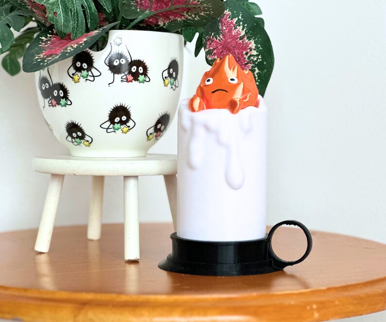 K&ouml;nnte beinhalten: Eine wei&szlig;e Kerze mit schwarzem Sockel und Griff. Die Kerze hat einen geschmolzenen Wachseffekt und eine kleine orangefarbene Figur auf der Oberseite. Die Figur ist ein Charakter aus dem Film "Mein Nachbar Totoro".