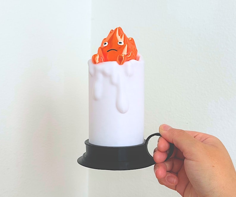 K&ouml;nnte beinhalten: Eine wei&szlig;e Kerze mit schwarzem Sockel und einer kleinen orangenen Figur eines Flammen auf der Oberseite. Die Figur hat einen traurigen Ausdruck.