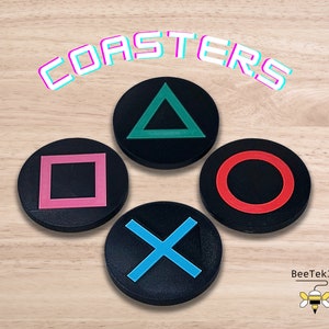 May include: Set of four black coasters with different colored geometric shapes: a green triangle, a pink square, a red circle, and a blue X. The coasters are labeled "COASTERS" in a pink and blue font.