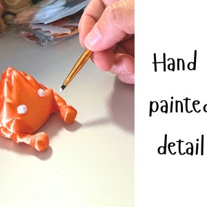 K&ouml;nnte beinhalten: Eine Hand bemalt Details auf einer kleinen, orangefarbenen 3D-gedruckten Krabbenfigur mit wei&szlig;en Augen. Handbemalte Details.