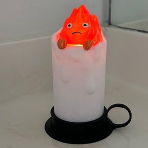K&ouml;nnte beinhalten: Eine wei&szlig;e Kerze mit schwarzem Sockel und einer roten und orangefarbenen Feuerspiritus-Figur auf der Oberseite. Die Figur hat einen traurigen Ausdruck und wird von innen beleuchtet.