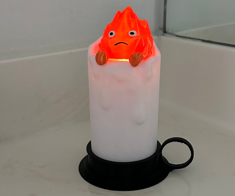 K&ouml;nnte beinhalten: Eine wei&szlig;e Kerze mit schwarzem Sockel und einer roten und orangefarbenen Feuerspiritus-Figur auf der Oberseite. Die Figur hat einen traurigen Ausdruck und wird von innen beleuchtet.