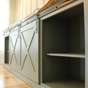 May include: A green cabinet with sliding barn doors. The doors have an X-shaped design and are made of wood. The cabinet has shelves inside and is made of wood. The cabinet is sitting on a wooden floor.