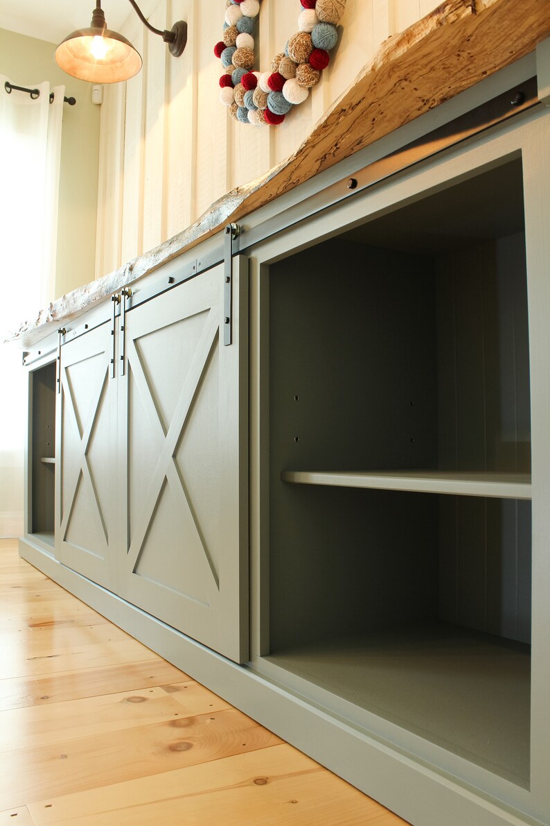 May include: A green cabinet with sliding barn doors. The doors have an X-shaped design and are made of wood. The cabinet has shelves inside and is made of wood. The cabinet is sitting on a wooden floor.