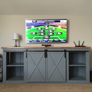 May include: A grey entertainment unit with two sliding barn doors, each with an X-shaped design. The doors are open, revealing shelves on either side. A flat-screen TV is mounted above the entertainment unit, displaying a football game lineup.