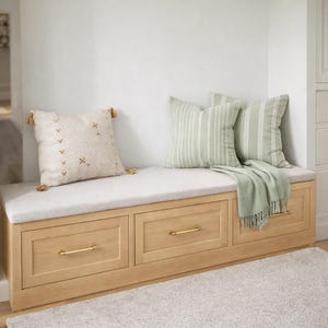 May include: A built-in wooden bench with three drawers, brass handles, and a light gray cushion. Two striped pillows and a textured pillow with tassels rest on the bench, along with a green throw blanket. The bench sits against a white wall.