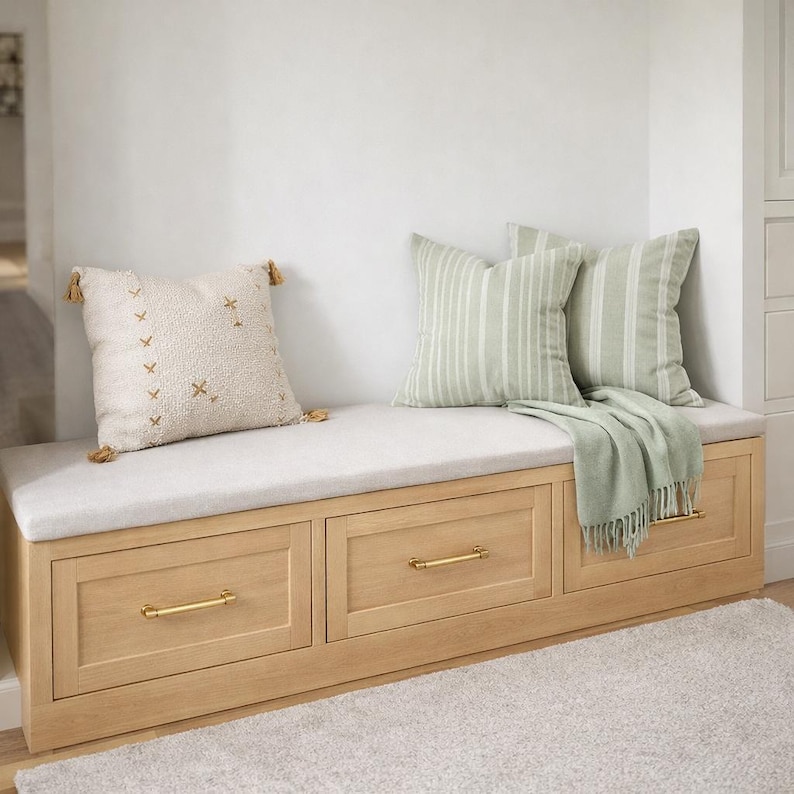 May include: A built-in wooden bench with three drawers, brass handles, and a light gray cushion. Two striped pillows and a textured pillow with tassels rest on the bench, along with a green throw blanket. The bench sits against a white wall.