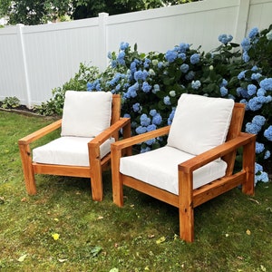 Puede incluir: Dos sillas de madera con cojines blancos se encuentran en un c&eacute;sped verde frente a una valla blanca y un arbusto de hortensias azules.