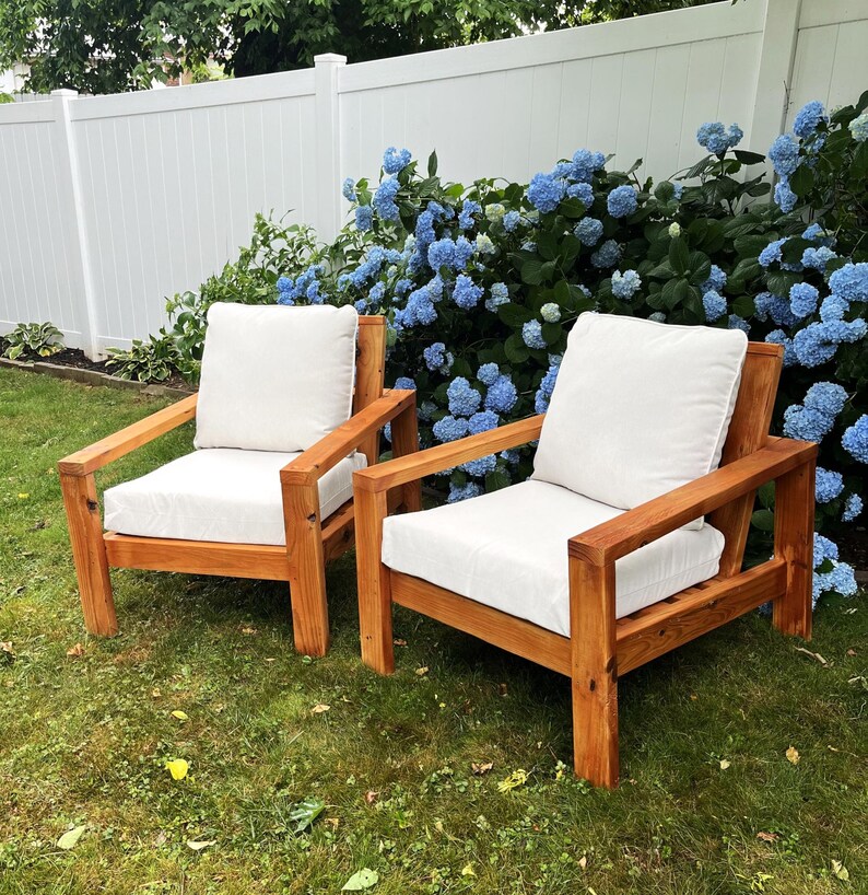 Puede incluir: Dos sillas de madera con cojines blancos se encuentran en un c&eacute;sped verde frente a una valla blanca y un arbusto de hortensias azules.