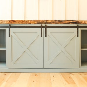 May include: A light blue media console with two sliding barn doors. The doors have an X-shaped design and black hardware. The console has two shelves on each side and a live edge wood top.