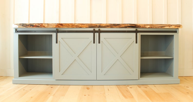 May include: A light blue media console with two sliding barn doors. The doors have an X-shaped design and black hardware. The console has two shelves on each side and a live edge wood top.