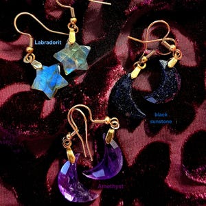 Könnte beinhalten: Drei Paar Ohrhänger mit goldfarbenen Haken. Ein Paar besteht aus sternförmigen Labradorit-Steinen, ein Paar aus halbmondförmigem schwarzem Sonnenstein und ein Paar aus halbmondförmigen Amethyst-Steinen, jeweils mit Beschriftung.