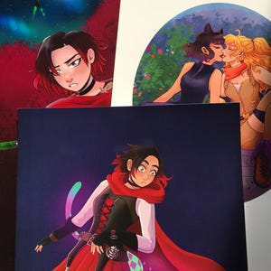 May include: Several anime-style art prints featuring characters with red and black hair, wearing capes and other fantasy attire. One print shows two characters kissing. The prints are on a table, with a dark blue background.