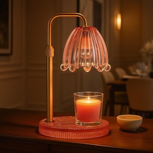 May include: A decorative candle warmer lamp with a pink glass shade and a gold-colored metal frame. The lamp sits on a textured pink base, next to a lit candle in a glass and a small wooden bowl. The lamp is designed to warm candles.