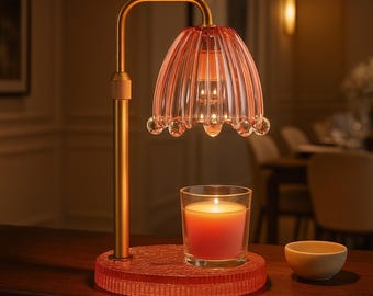 May include: A decorative candle warmer lamp with a pink glass shade and a gold-colored metal frame. The lamp sits on a textured pink base, next to a lit candle in a glass and a small wooden bowl. The lamp is designed to warm candles.
