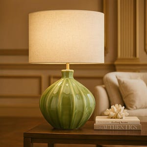 May include: A green ceramic table lamp with a beige linen shade. The lamp base has a ribbed design. The lamp is on a dark brown wooden table. The lamp is a decorative home decor item.