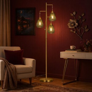 May include: A gold-toned floor lamp with three green glass shades. The lamp has a tall, slender pole with a round base and a branching top. The shades are a textured, ribbed design, and the lamp is placed in a room with a dark red wall.