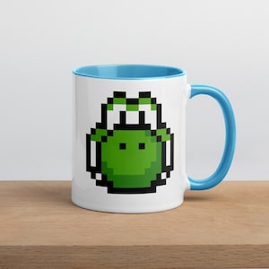 May include: White ceramic mug with a pixelated green Yoshi design. The interior and handle are light blue. The mug is on a wooden surface.