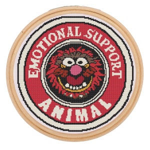 Op de afbeelding: Een ronde borduurring in rood en wit met een zwart-wit afbeelding van een roodharige, roze-neus monster met de tekst "Emotional Support Animal" rond de afbeelding.