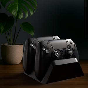 Op de afbeelding: Zwarte gamecontrollers op een zwart oplaadstation, geplaatst op een houten bureau. Een computermonitor, toetsenbord en potplant zijn ook zichtbaar. De controllers hebben meerdere knoppen en joysticks.