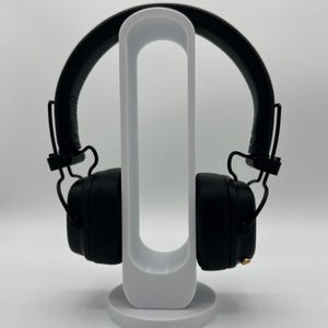Pu&ograve; includere: Cuffie over-ear nere con archetto imbottito e padiglioni quadrati, dotate di una piccola manopola di controllo dorata sul lato destro. Le cuffie sono appoggiate su un supporto espositivo bianco, verticale e di forma ovale con base circolare.