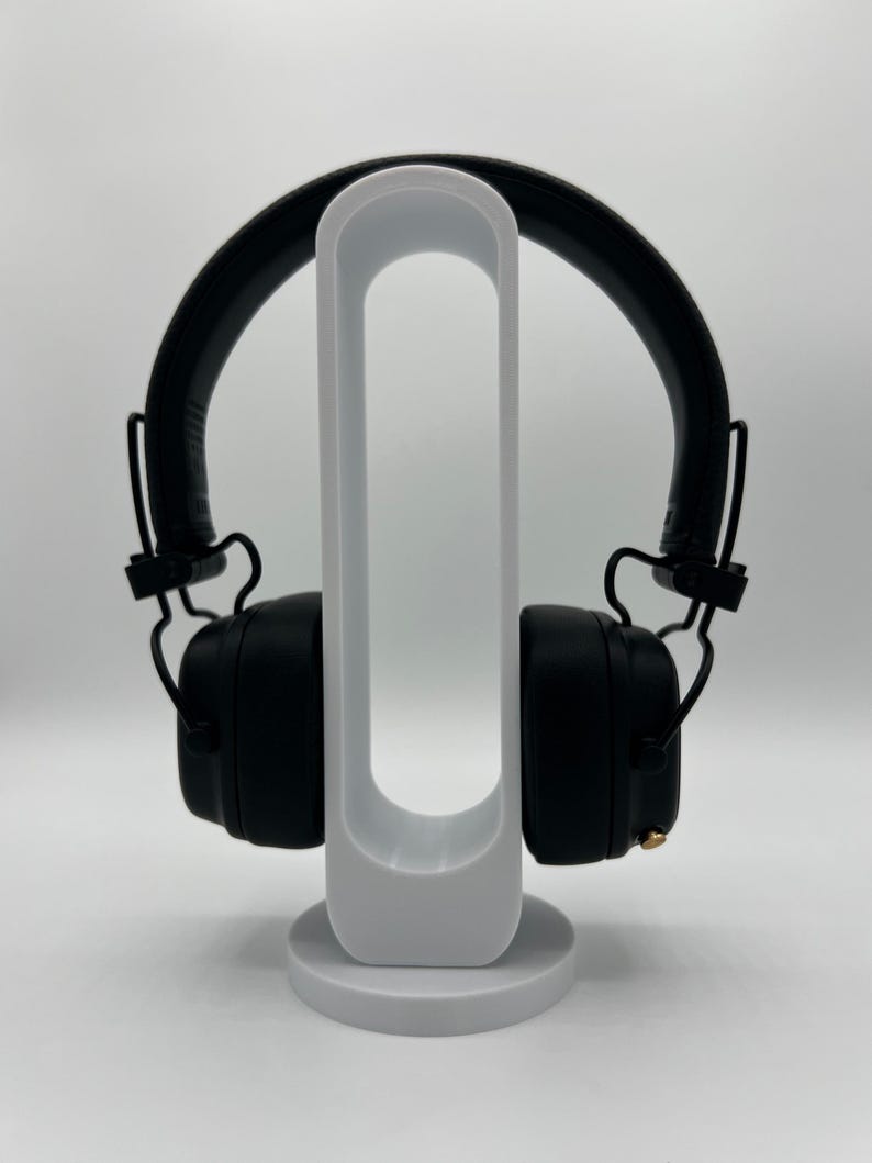 Pu&ograve; includere: Cuffie over-ear nere con archetto imbottito e padiglioni quadrati, dotate di una piccola manopola di controllo dorata sul lato destro. Le cuffie sono appoggiate su un supporto espositivo bianco, verticale e di forma ovale con base circolare.