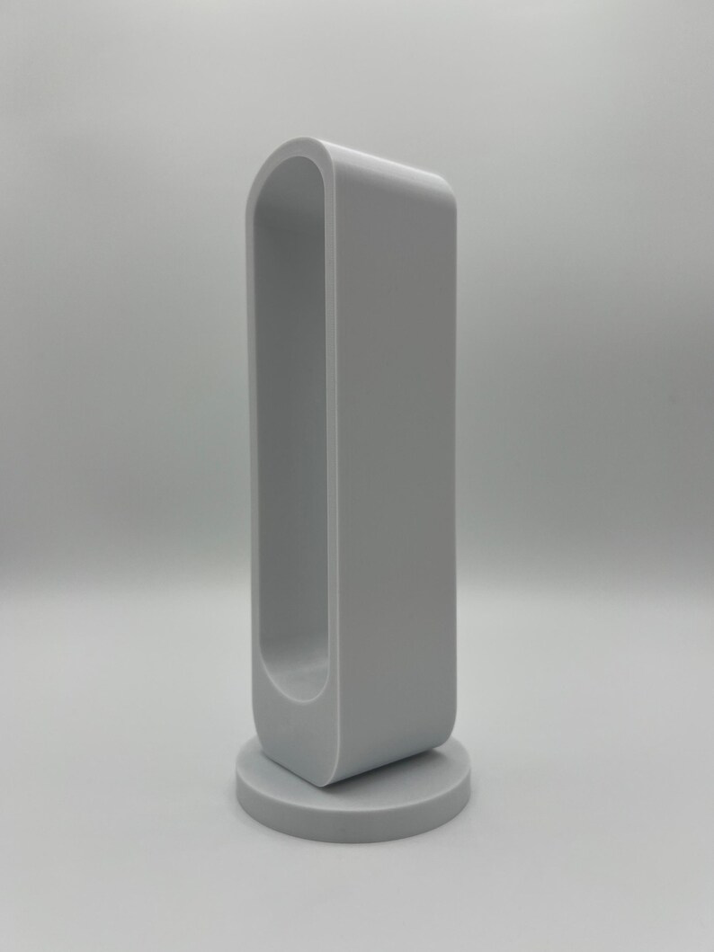 Pu&ograve; includere: Un oggetto grigio chiaro, orientato verticalmente, di forma rettangolare cava con angoli superiori arrotondati, montato su una base circolare. La struttura presenta una cornice continua e liscia con un'ampia apertura allungata al centro.