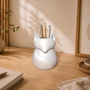 Könnte beinhalten: Ein mattweißer Schreibtisch-Organizer in Fuchsform mit einer glatten, abgerundeten Silhouette. Der Fuchs ist in einer zusammengerollten Ruheposition mit geschlossenen Augen und einem um die Seite gewickelten Schwanz dargestellt. Drei Stifte stecken aufrecht.