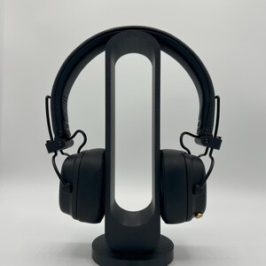 Pu&ograve; includere: Cuffie nere over-ear appoggiate su un supporto verticale nero di forma ovale. Le cuffie presentano un archetto testurizzato, padiglioni imbottiti e una piccola manopola di controllo color oro sul lato destro. Il supporto ha una base circolare.