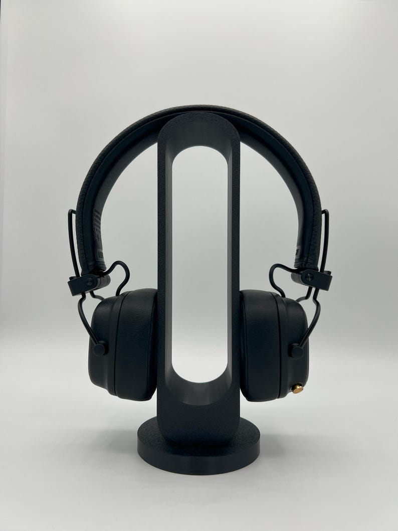Pu&ograve; includere: Cuffie nere over-ear appoggiate su un supporto verticale nero di forma ovale. Le cuffie presentano un archetto testurizzato, padiglioni imbottiti e una piccola manopola di controllo color oro sul lato destro. Il supporto ha una base circolare.